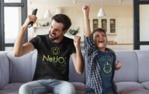 Father and son sitting in their living room in NetJOI branded shirts - son's hand is raised to the ceiling, Dad's hands are pumped.