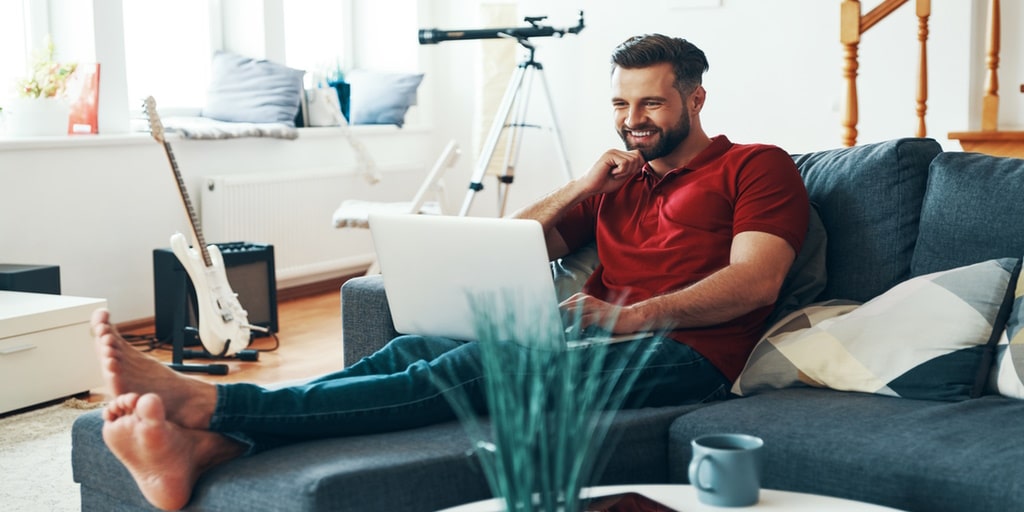 Best, Reliable and Most Affordable Internet Providers in Ontario for Under $50 per Month - Young man in casual clothing using laptop and smiling while sitting on the sofa.
