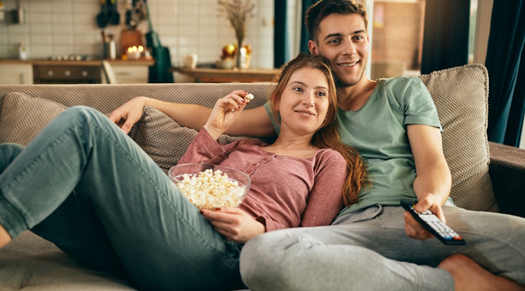 Smiling couple, eating popcorn, sitting in sofa watching NetJOI TV.
